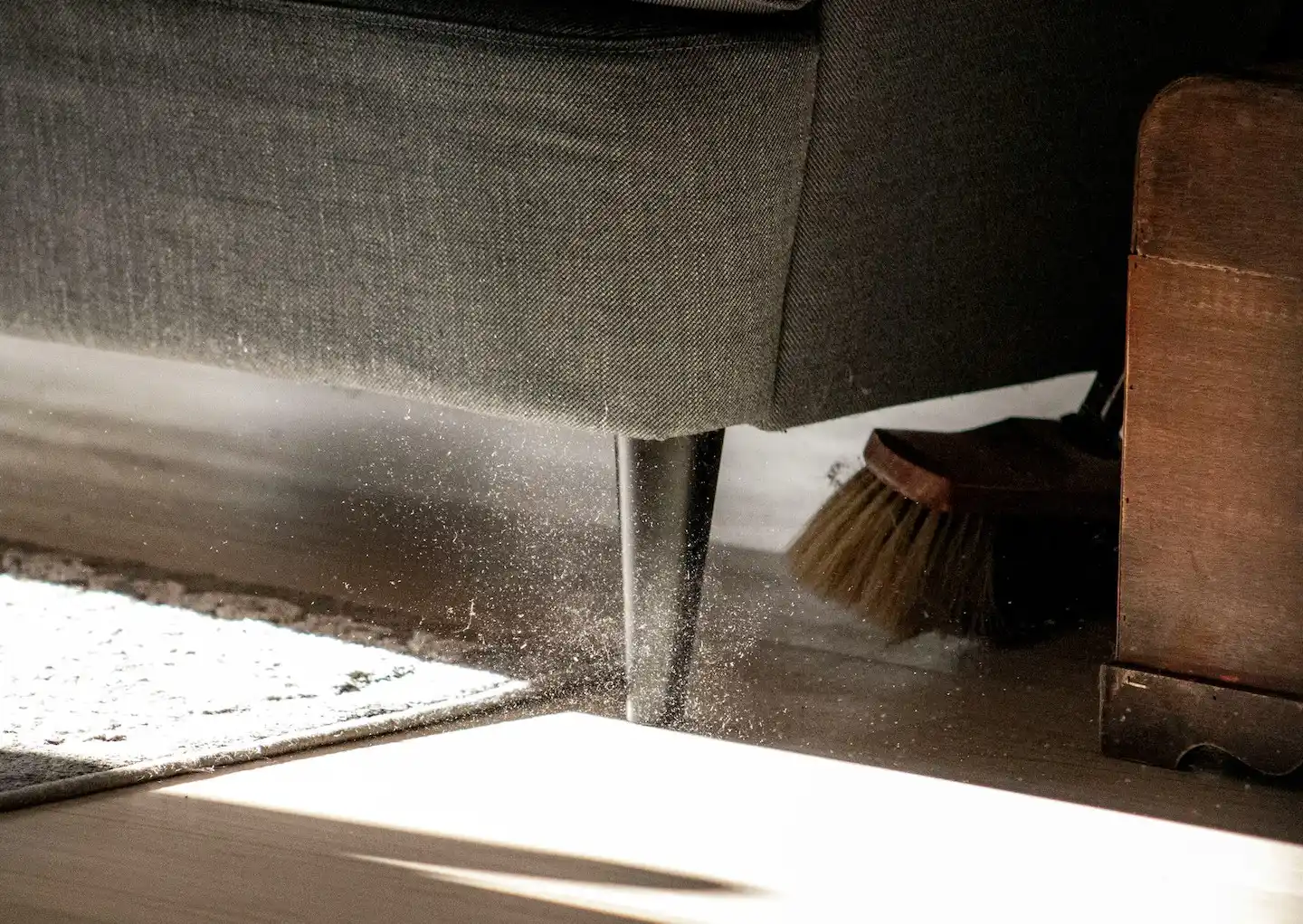 Person sweeping under couch as dust flies into the air.
