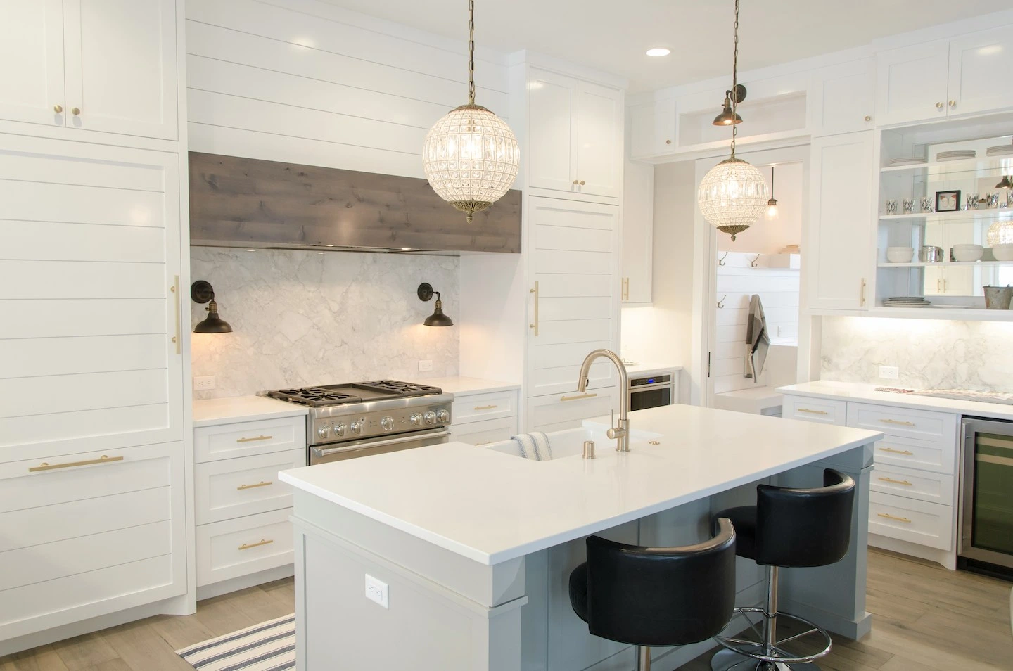 A white island table with two black chairs in the center of a white painted kitchen, and two glass lamps hanging over the island. 